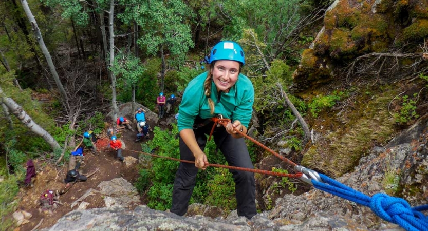 From above, a person wearing safety gear smiles as they climb towards the camera. There are people on the ground below. 